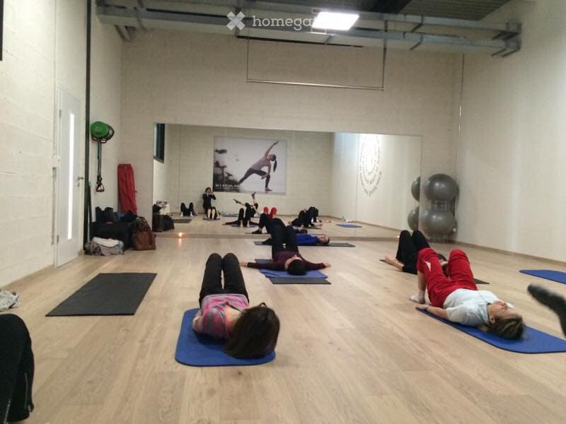A spacious studio with wooden flooring, several yoga mats laid out, and a few fitness balls in the corner. A poster of a yoga pose is displayed on the wall, and bags are placed near the entrance.