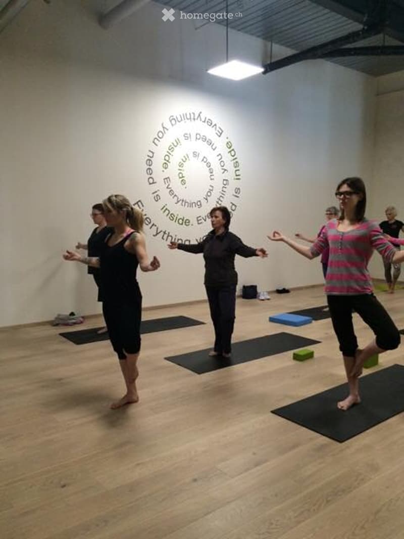 A yoga studio with wooden flooring and white walls. Several people are practicing yoga on black mats. A blue yoga block and a green yoga block are visible on the floor.