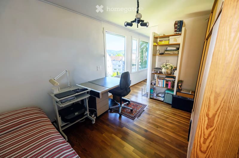A well-lit room with wooden flooring, a desk with a laptop and printer, a bookshelf filled with books and decorative items, and a comfortable chair. The room features large windows with a scenic view and a cozy rug on the floor.