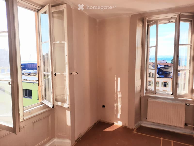 Empty room with light-colored walls, wooden floor, and large windows offering a view of the city and sea.