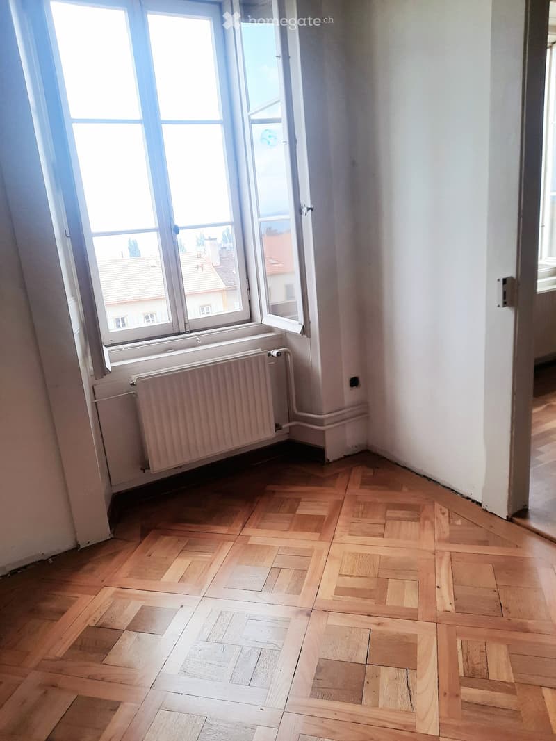 Empty room with wooden parquet flooring, white walls, large window with radiator below, and a view of rooftops outside.