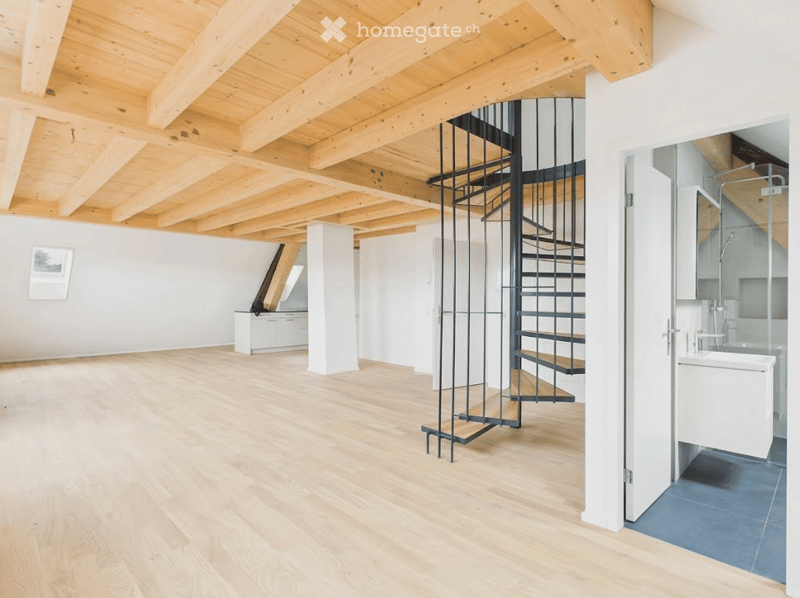 Open-plan space with exposed wooden beams, light wooden flooring, a modern spiral staircase, and a small bathroom area with a bathtub and glass shower enclosure.