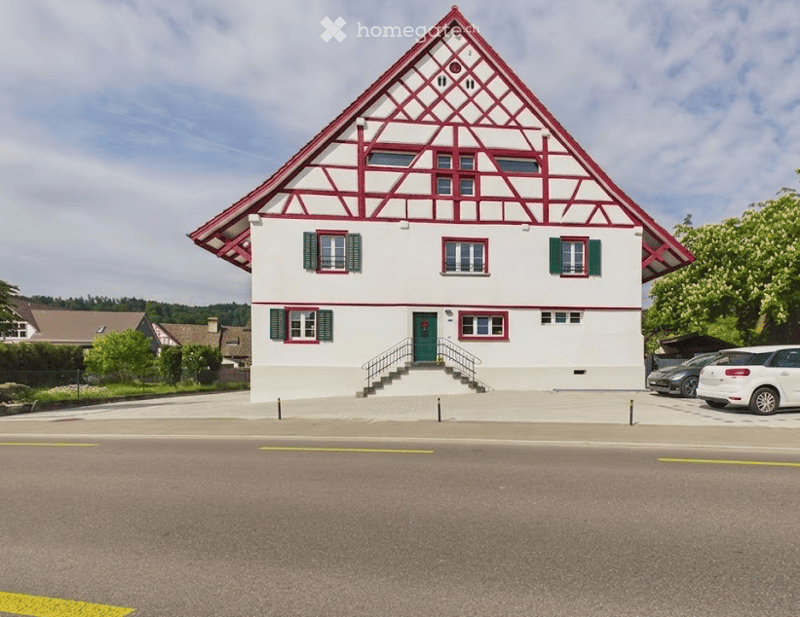 Two-story house with white walls, green shutters, red trim, and a distinctive red triangular roof. A small staircase leads to the front door, and there are parked cars in the driveway.