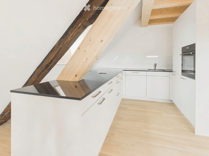 Modern kitchen with white cabinets, black countertop, and stainless steel appliances, featuring exposed wooden beams and a wooden floor.