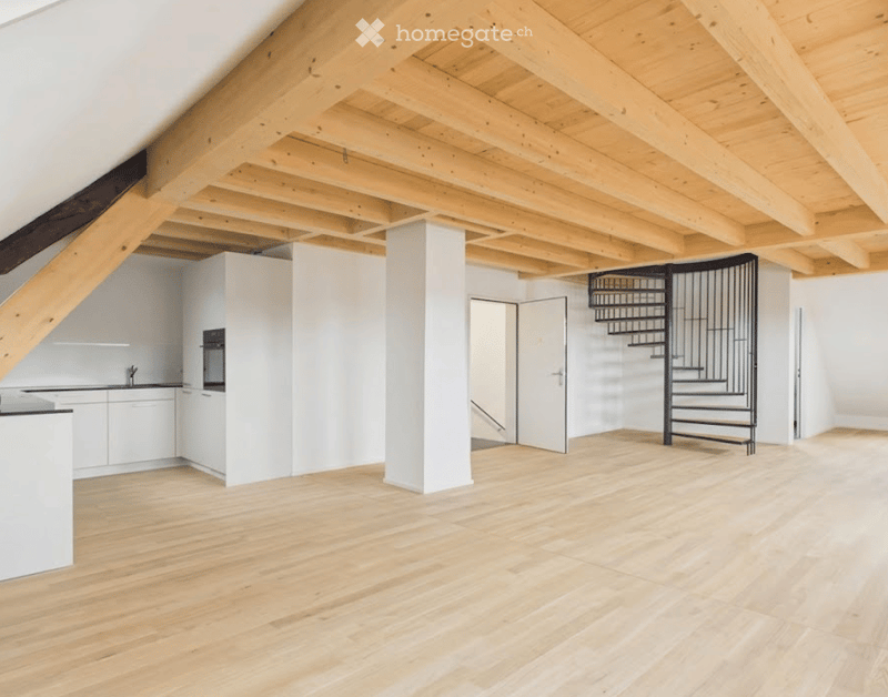 Bright, open loft space with wooden floors and exposed wooden beams. Features a modern white kitchen area with cabinets and a sink, a black spiral staircase leading to an upper level, and large white walls.