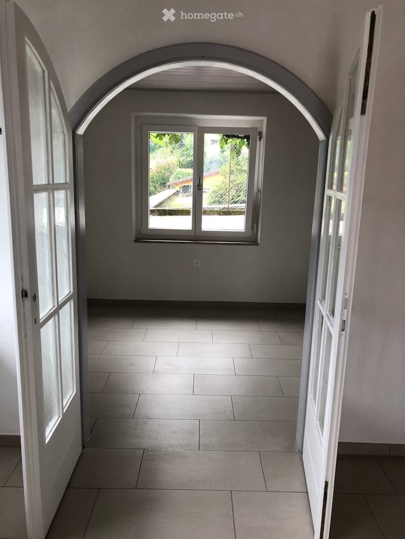 Empty room with arched entrance, white tiled floor, white walls, large window overlooking a garden.