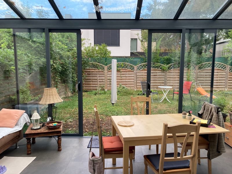 A well-lit conservatory with a wooden dining table and chairs, a sofa with a pink cushion, a lamp, and a small table with a decorative lantern and a bowl. The conservatory has large glass walls and doors opening to a garden with a wooden fence, outdoor fu