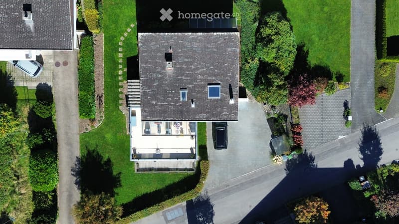 Aerial view of a two-story house with a grey roof, multiple windows, and a well-maintained garden. A driveway leads to a garage with a parked car, and there is a patio area with outdoor furniture.