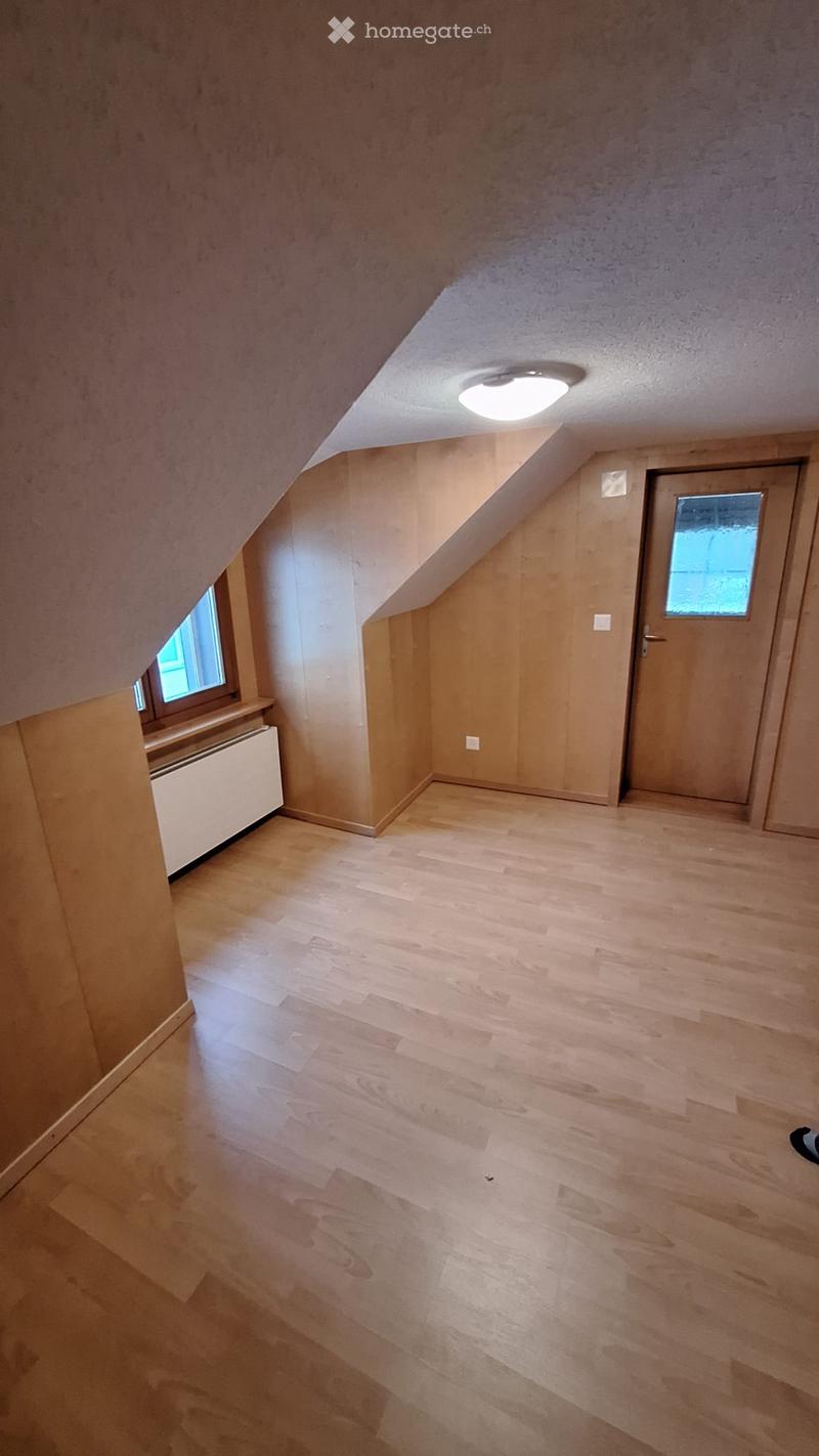 Empty attic room with wooden flooring, slanted ceilings, and a single door with a window. Natural light from small windows illuminates the space.