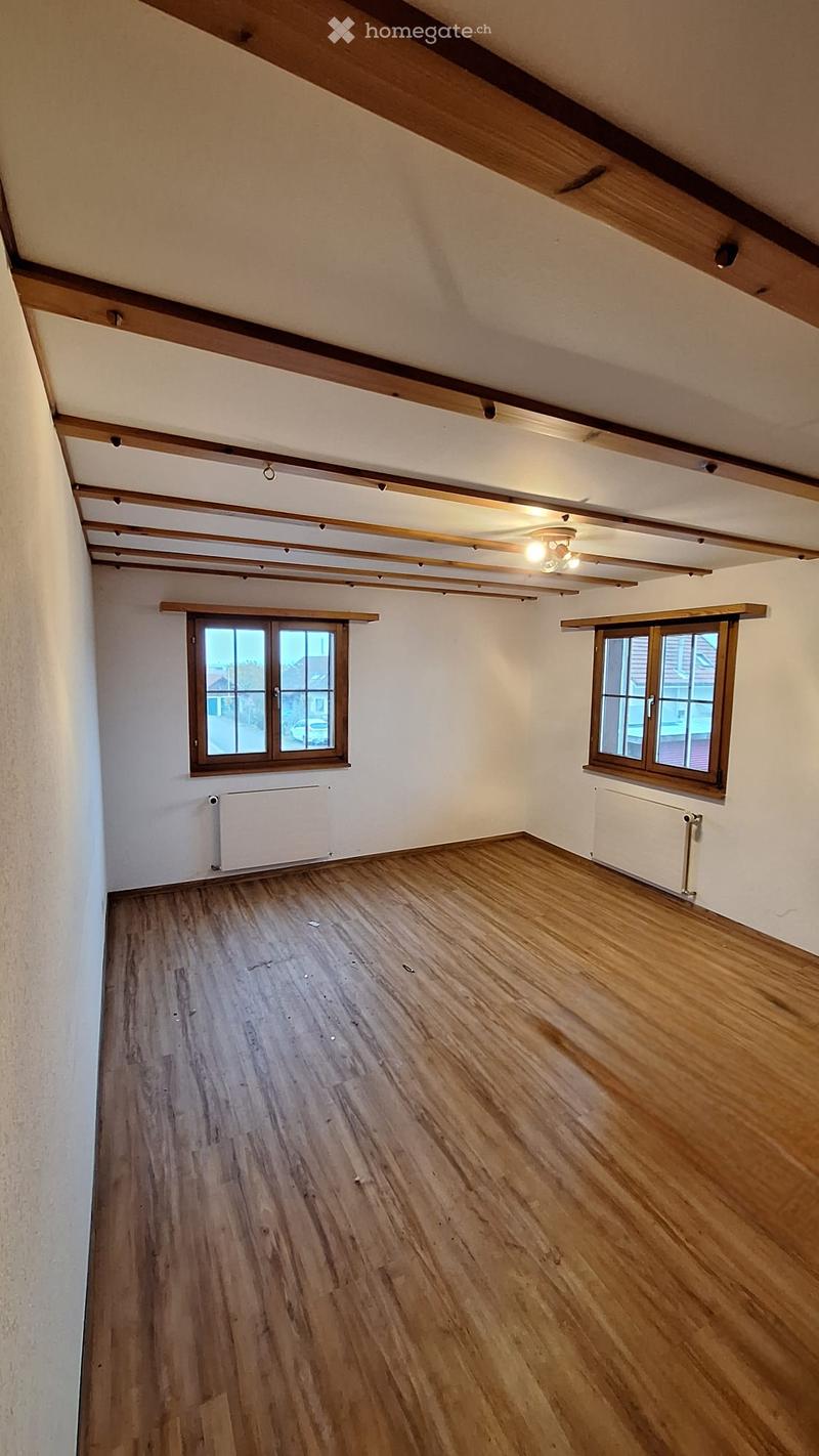 Empty room with wooden flooring, white walls, exposed wooden beams on the ceiling, two windows with wooden frames, and radiators on the walls.