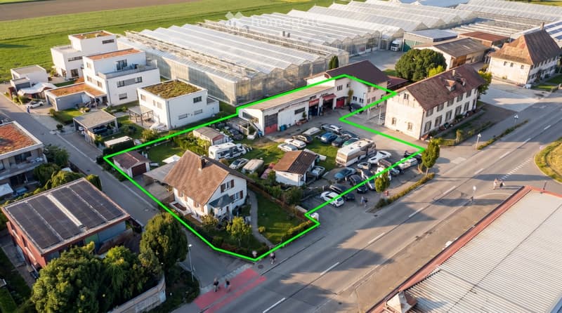 Aerial view of a residential and commercial area with multiple buildings, parking lots, greenhouses, and surrounding greenery.