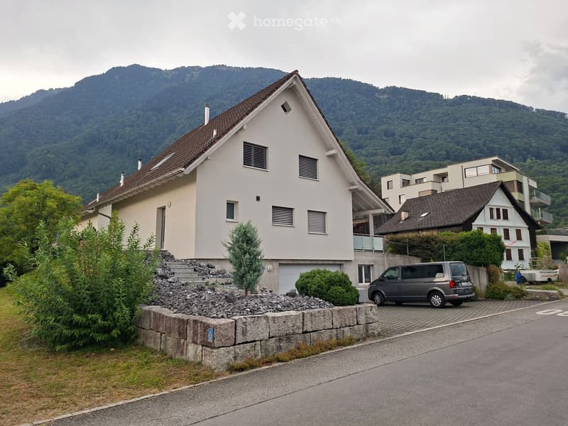 Two-story house with a brown roof, white walls, and multiple windows, situated in a scenic area with mountains in the background. A grey van is parked in the driveway.