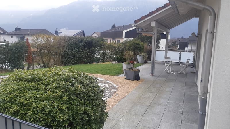 Balcony with tiled floor, table and chairs, surrounded by greenery and a view of the mountains.