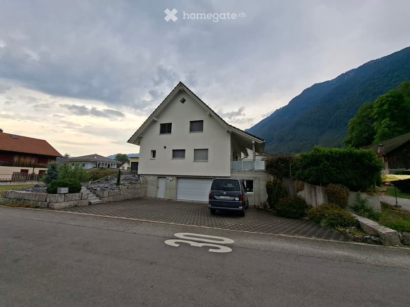 Two-story house with white exterior, garage, and balcony, situated in a scenic mountainous area with surrounding greenery.