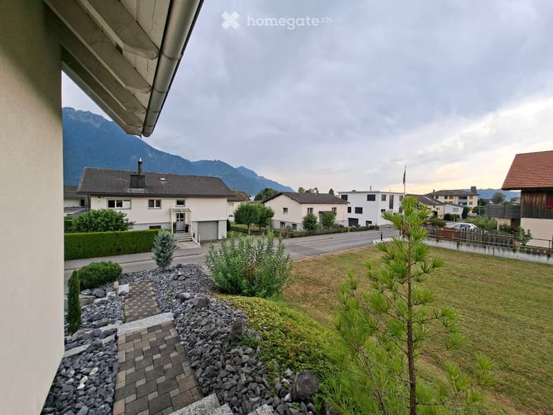 View from a balcony showing a well-maintained garden with grass, small trees, shrubs, and a stone pathway leading to a house entrance. The background features a residential street with houses and distant mountains.