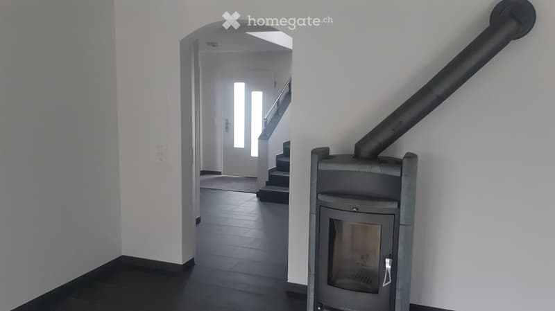 Modern hallway with a sleek black tiled floor, white walls, and a visible staircase leading upwards. A contemporary wood-burning stove is positioned on the right side.