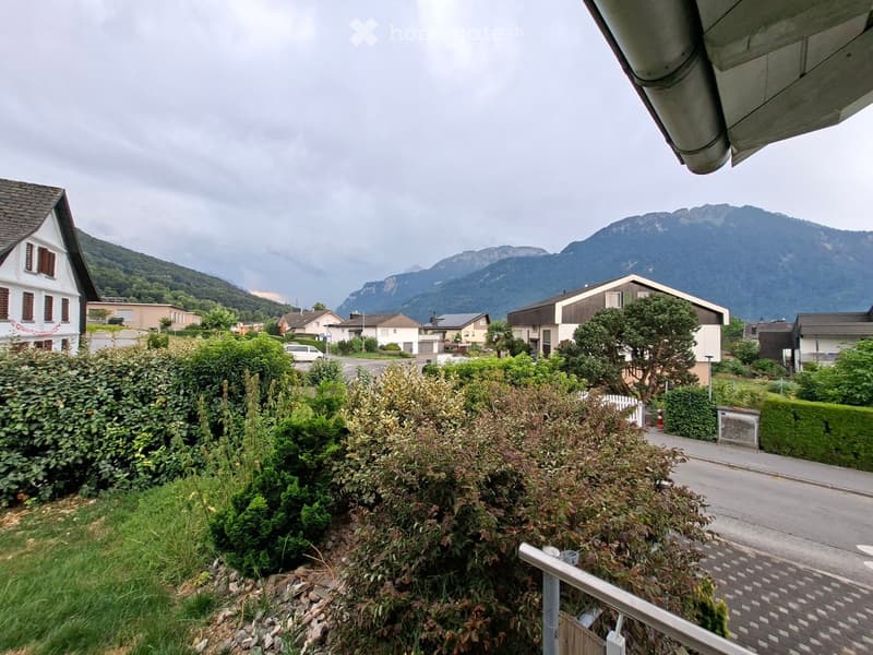 View from a balcony or terrace showing a suburban neighborhood with houses, greenery, and distant mountains.