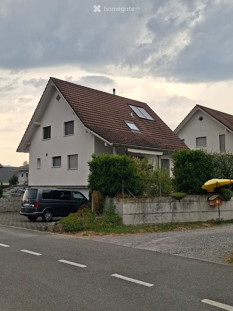 Two-story house with a brown tiled roof, white walls, multiple windows, and a small garden area in front.
