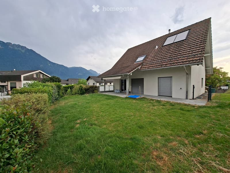 Single-family house with a brown tiled roof, two garage doors, and a well-maintained lawn in front. The house is situated in a residential area with neighboring houses and a scenic mountain backdrop.