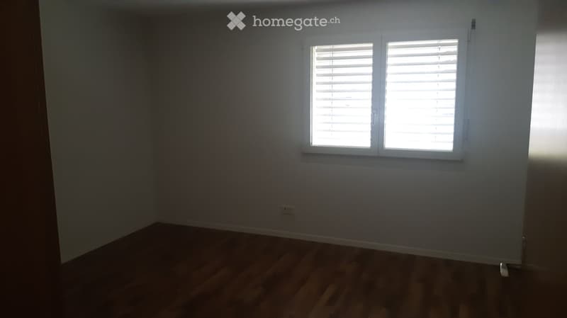 Empty room with wooden flooring and white walls, featuring a window with white blinds and a visible electrical outlet on the wall.