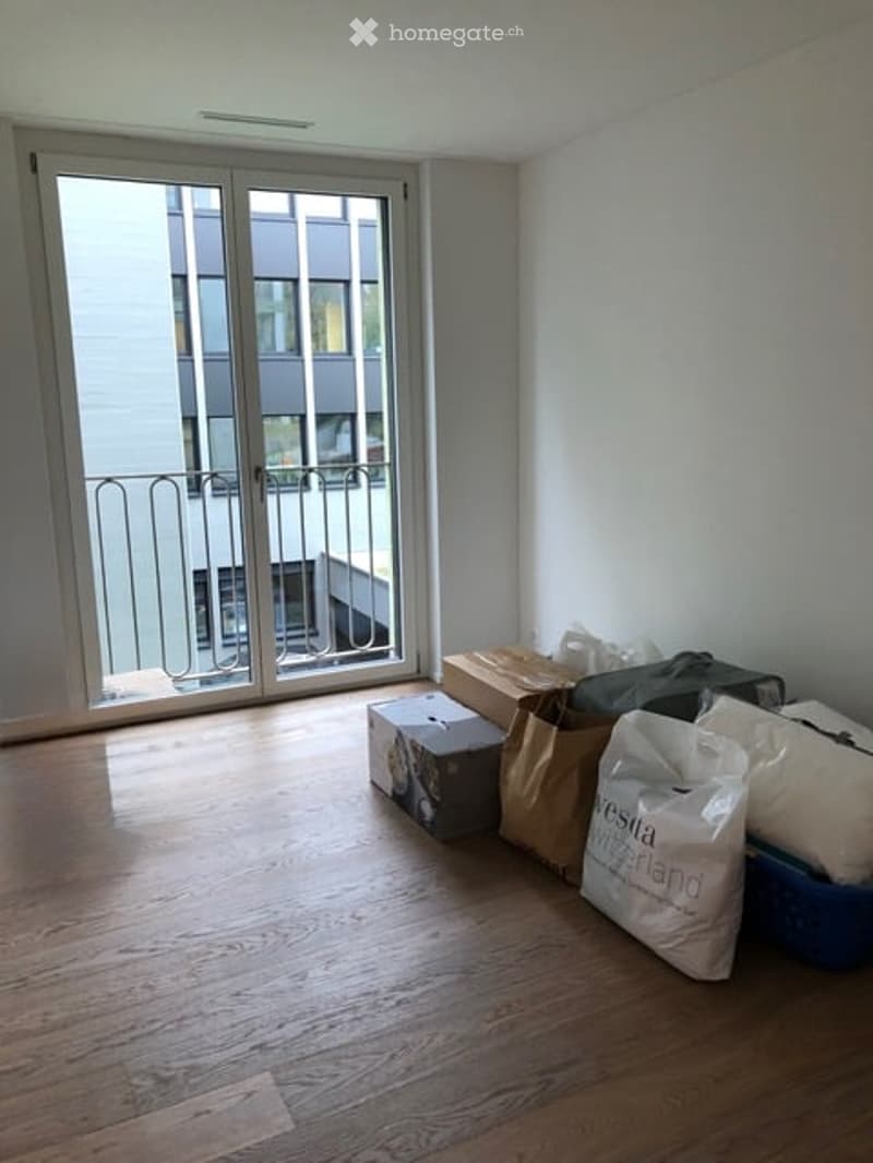 Empty room with wooden flooring, large sliding glass door leading to a balcony, and several cardboard boxes and bags placed near the door.