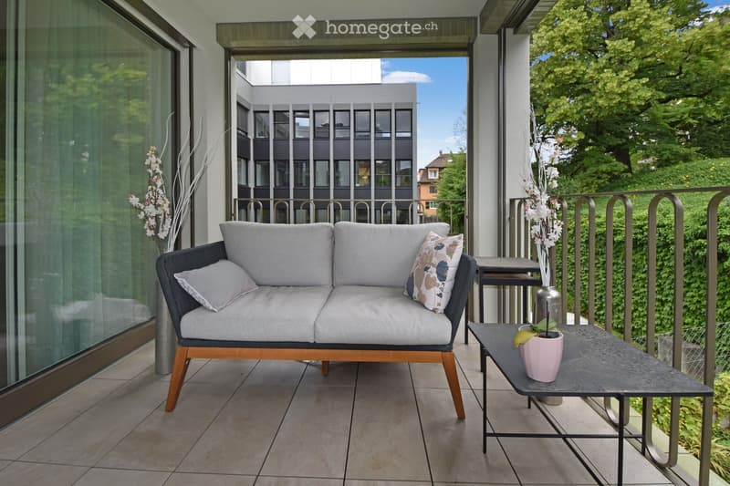 Balcony with a grey sofa, small table, and decorative vases, overlooking a green area with trees and a modern building.