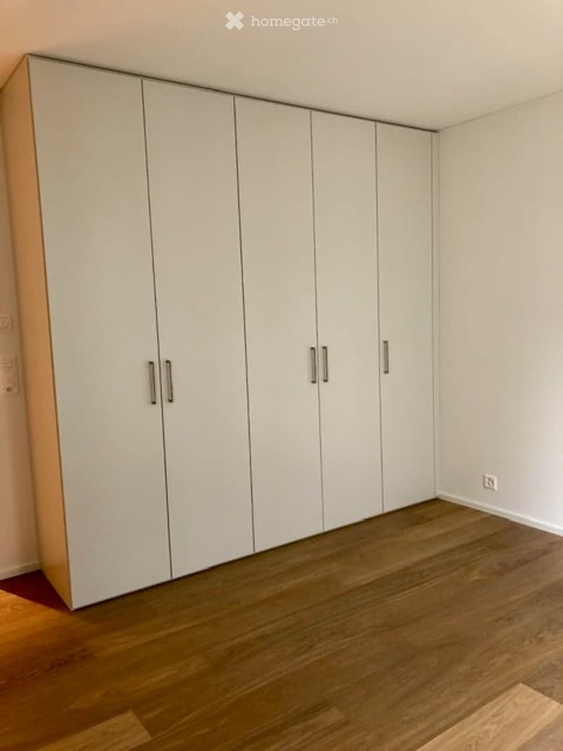 Empty room with light-colored wooden flooring and white built-in wardrobes along one wall.