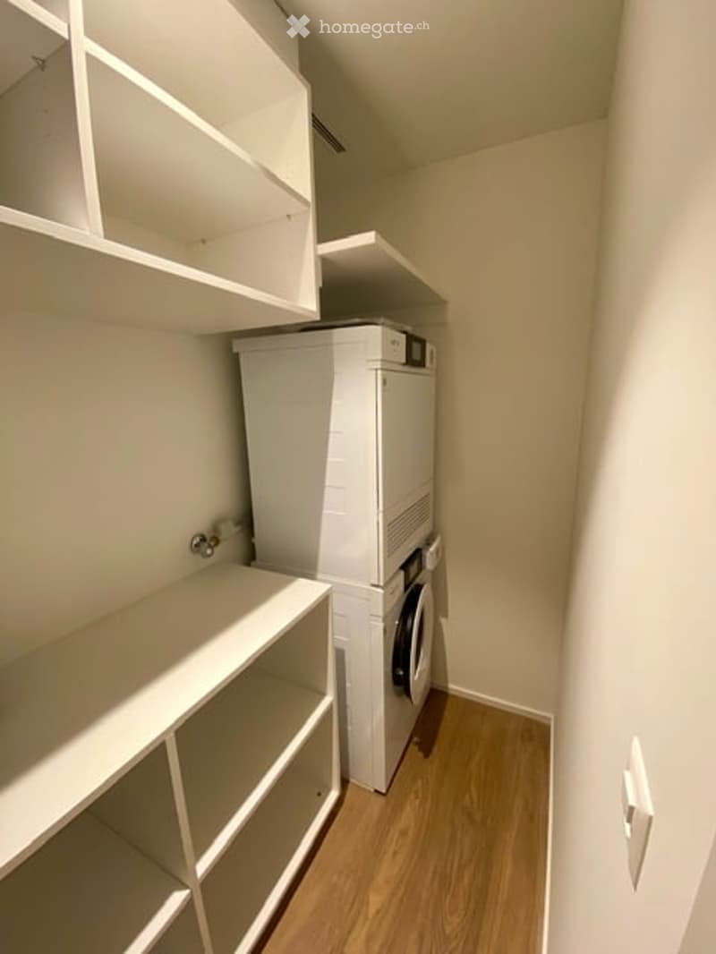 A small, well-lit laundry room with built-in white shelves and a white washing machine. The floor is light wood, and the walls are painted a neutral beige.
