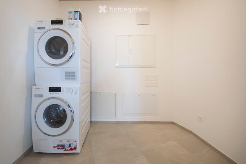 A small, clean room with a stacked washing machine and dryer unit against one wall, white tiled floor, and white walls with built-in cabinets.