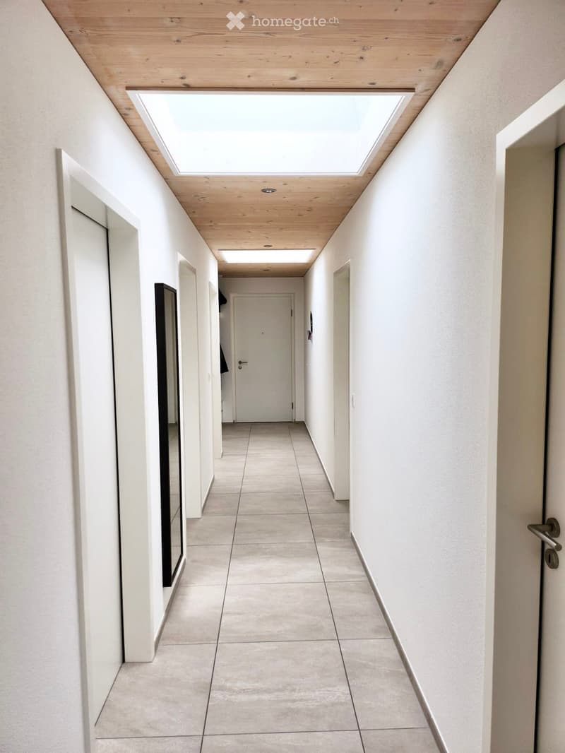 Modern hallway with light wooden ceiling, skylights, tiled floor, and white walls. Features several doors and a mirror on one side.