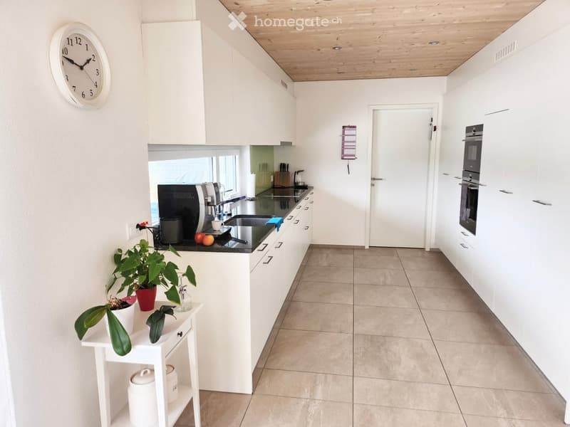 Modern kitchen with white cabinets, stainless steel appliances, and a black countertop. Features include a coffee machine, sink, oven, fridge, and a small table with plants and a clock on the wall.