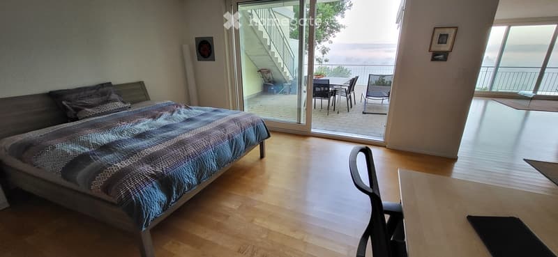 Bright bedroom with wooden flooring, large bed with patterned bedding, a chair, and a table near the sliding glass doors leading to a balcony with outdoor furniture and a scenic view.