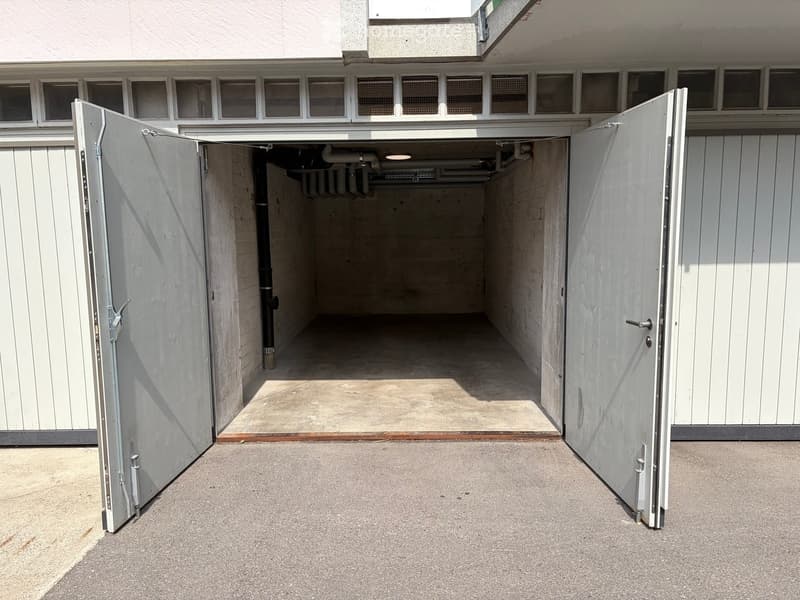 Open garage with large, white, sliding doors, concrete floor, and visible pipes and lighting inside.