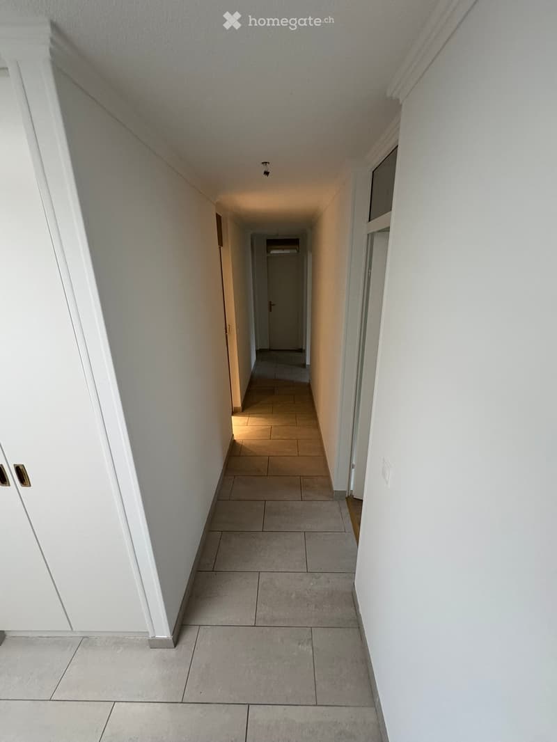 Empty hallway with tiled floor and white walls, featuring doors on both sides and a small window at the far end.