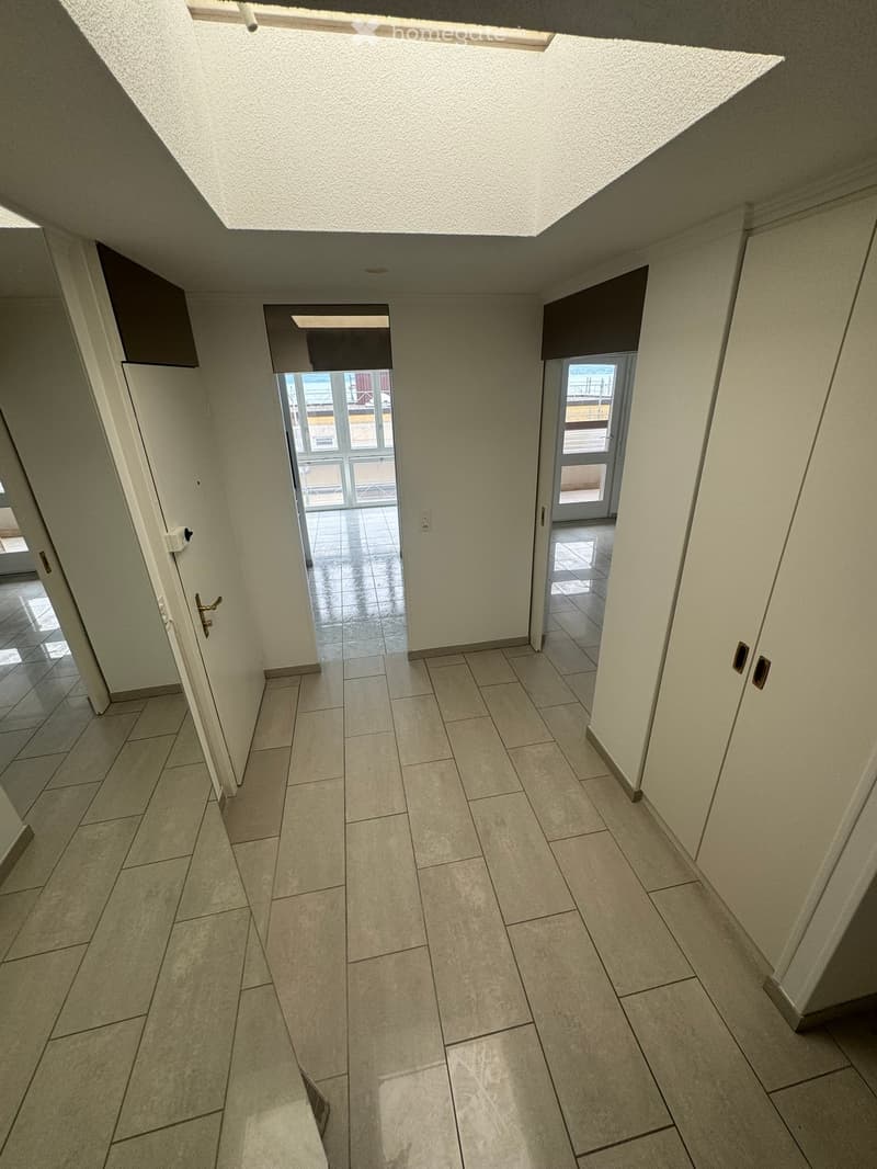 Empty hallway with tiled flooring, white walls, and multiple doors leading to other rooms. Natural light streams in from the end of the corridor.