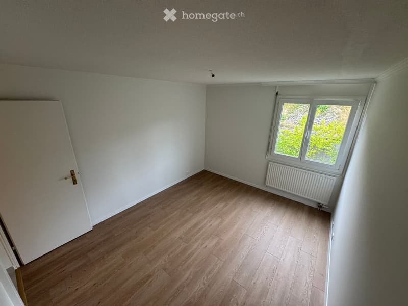 Empty room with wooden flooring, white walls, a window with a view of greenery, and a radiator below the window.