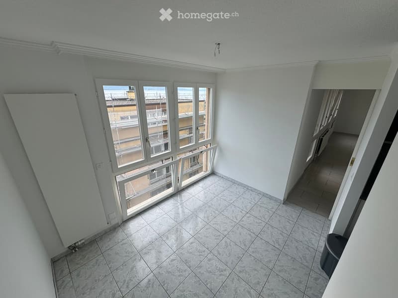 Empty room with tiled floor, white walls, large windows with white frames, and a small trash bin in the corner.