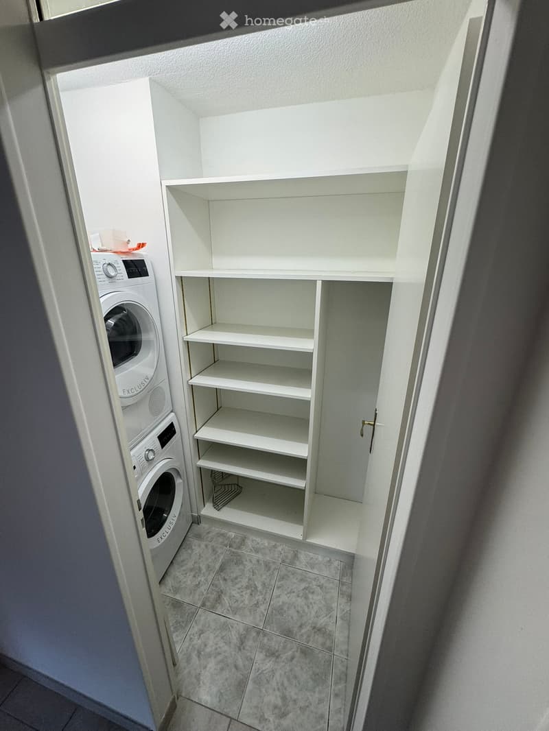 A small, well-lit laundry room with a white washing machine and a built-in white cabinet with multiple shelves.