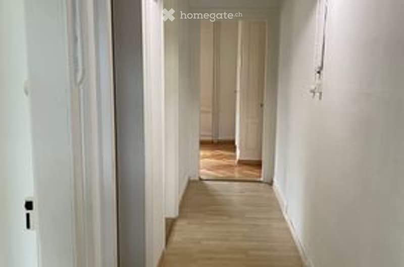Empty hallway with light-colored walls and wooden flooring, leading to an open doorway at the end.