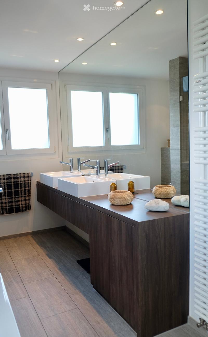 Modern bathroom with a double sink vanity, sleek wooden cabinetry, and a large mirror above the sinks. The countertop features decorative items including wicker baskets and stones.