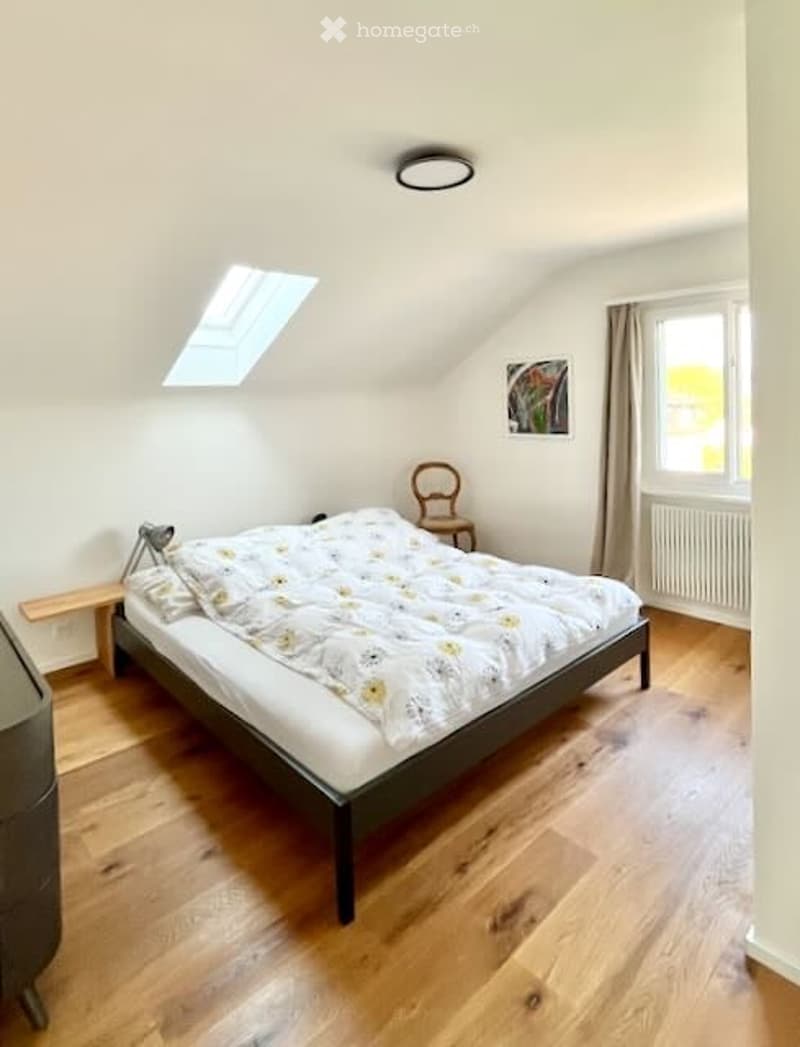 Cozy attic bedroom with wooden flooring, a double bed with a floral duvet, a wooden chair, a picture frame on the wall, and a skylight window.