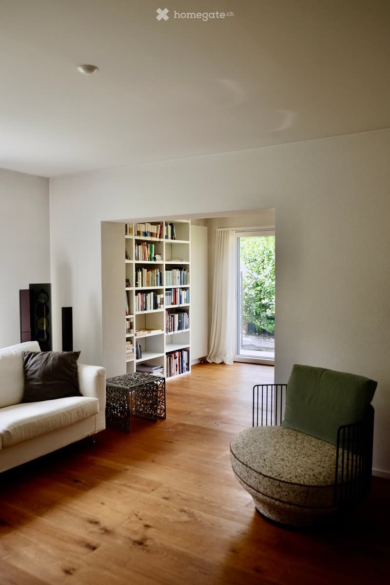 Living room with wooden flooring, white sofa, green chair, small table, bookshelf, and speakers.