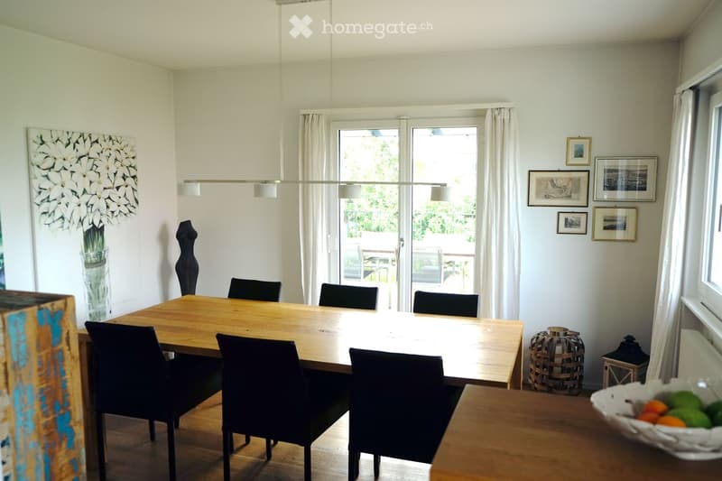 Bright dining room with a wooden dining table surrounded by black chairs, a vase with white flowers, framed pictures on the wall, and a wicker basket near the window.