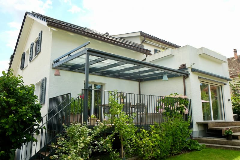 Two-story house with a covered patio area, black metal railing, and potted plants. The house has white walls, multiple windows, and a staircase leading to the patio.