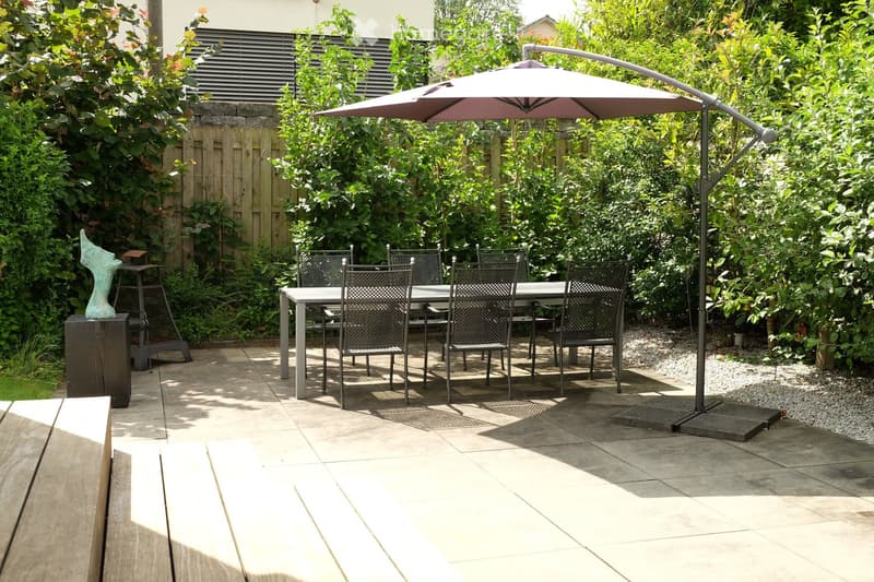 Garden patio with a round table, six chairs, and a large umbrella providing shade. A decorative sculpture sits on a wooden stand to the left. The area is surrounded by lush greenery and a wooden fence.