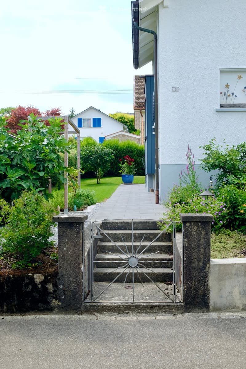 A well-maintained garden with a metal gate leading to a house entrance, surrounded by lush greenery and flowers. The pathway is paved, and the house features a white facade with blue shutters and a decorative window.