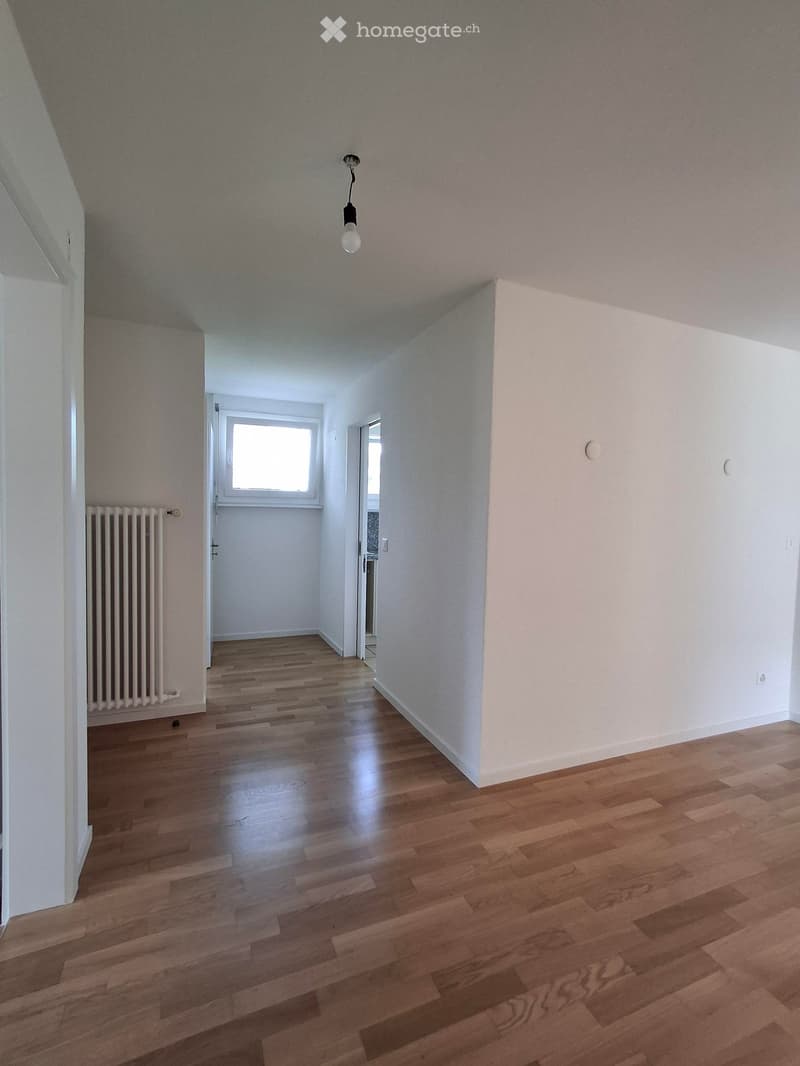 Empty room with wooden flooring, white walls, and a radiator on the left side. A small window and a door are visible in the background.