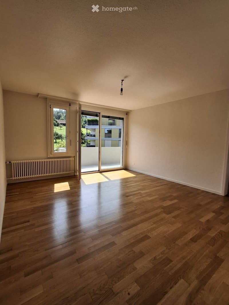 Empty room with wooden flooring, white walls, large window, radiator, and sliding glass door leading to a balcony.