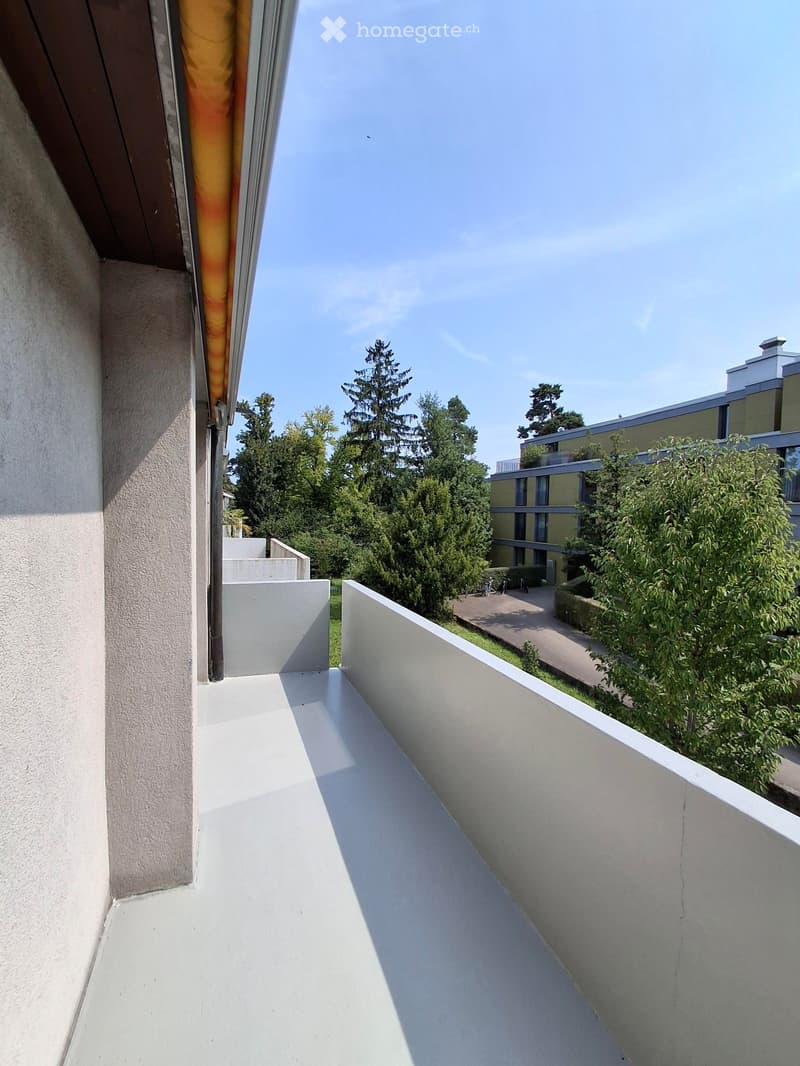 Balcony with white walls and a clear view of a landscaped area with trees and a modern building in the background.