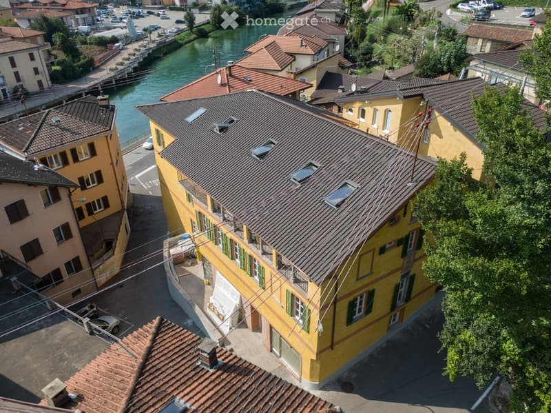 Aerial view of a multi-story yellow building with a tiled roof, situated near a river with surrounding residential structures and parked cars.